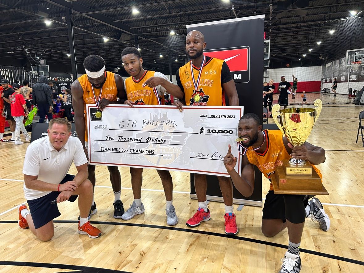 GTA Ballers team in orange jerseys holding an oversized $10,000 cheque for Team Nike 3vs3 Champions dated July 29th 2023 and the Team Nike Cup trophy on an indoor court